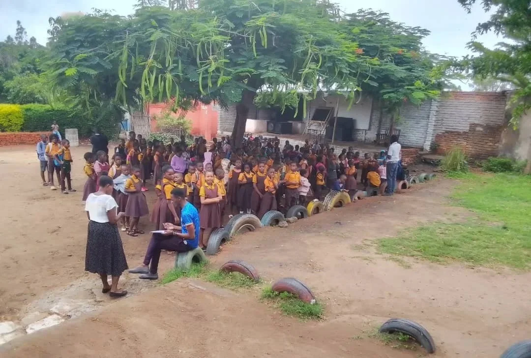 Schulhof mit mehreren dutzend Schulkindern in Uniformen, außerdem ein paar Erwachsene in Straßenklamotten. Auf dem Schulhof stehen Bäume.