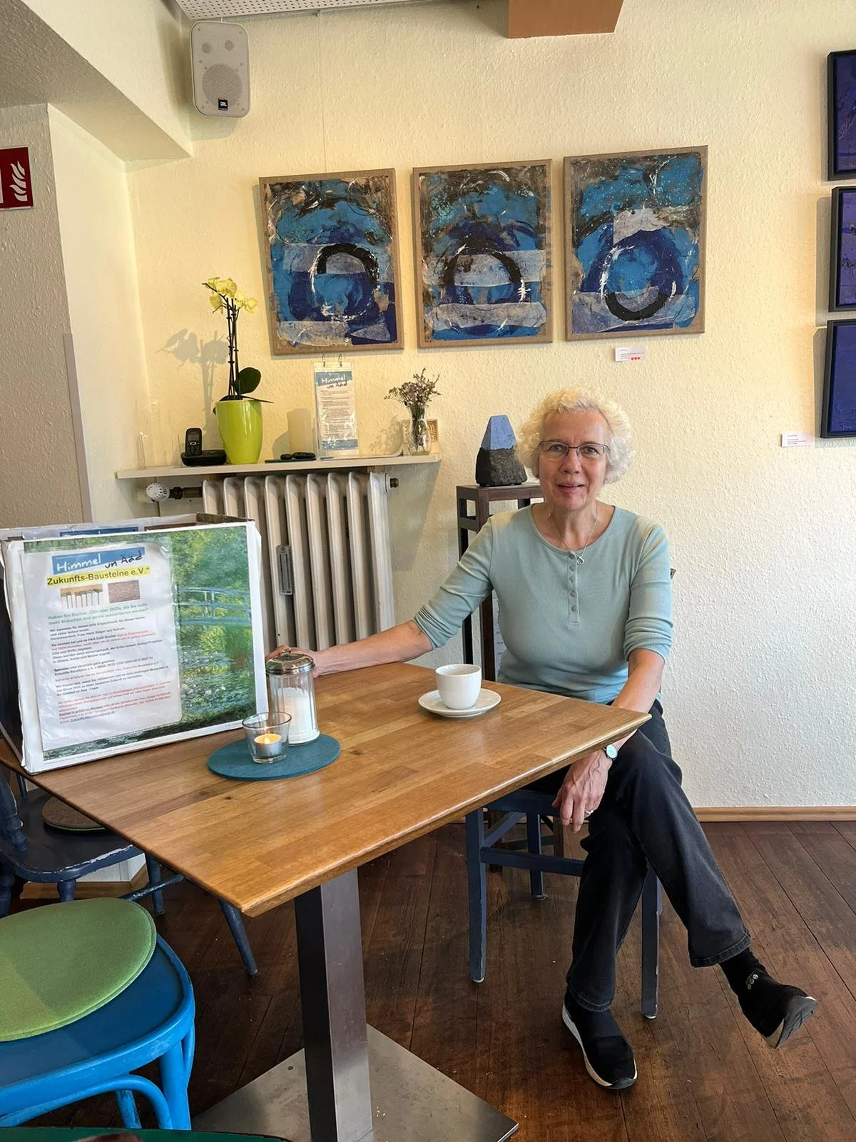 Ältere Frau sitzt an einem Tisch in einem Café, auf dem Tisch steht ein Bilderrahmen mit einem Text zum Verein Zukunfts-Bausteine