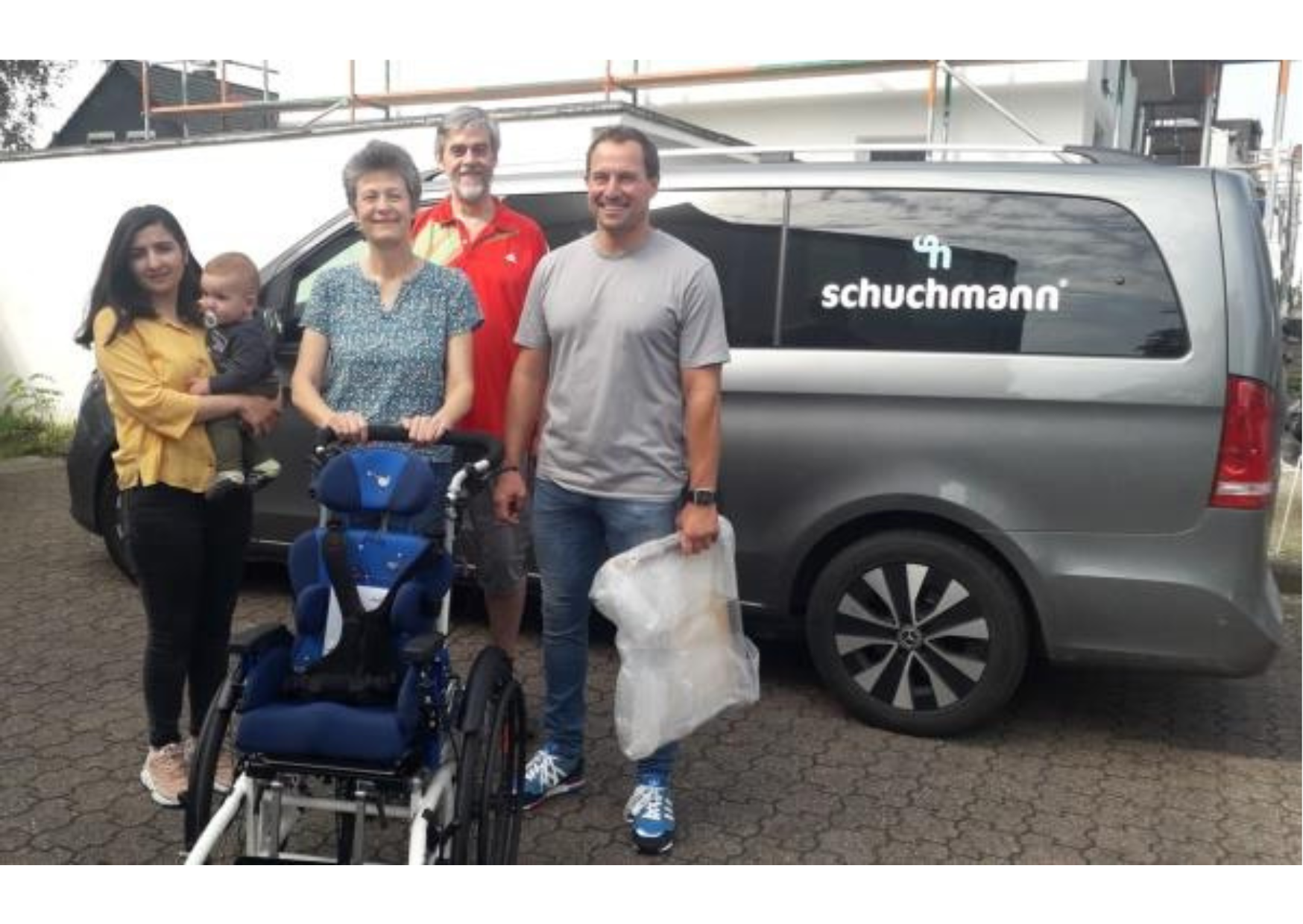 Eine Frau mit Baby auf dem Arm, ein Pärchen um die 60 Jahre alt und ein Mann um die 45 Jahre alt stehen nebeneinander. Vor ihnen ein blauer Therapiestuhl. Hinter ihnen ein silberner Transporter mit der Aufschrift schuchmann. Alle Personen lächeln.