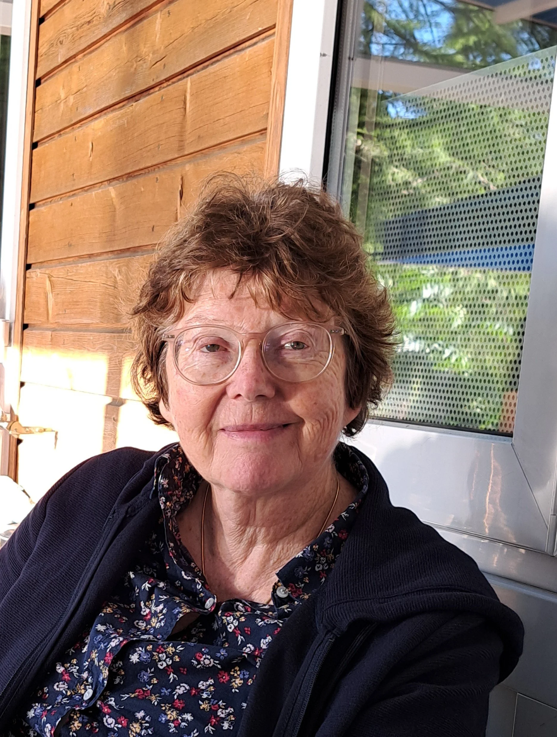 Nahaufnahme einer älteren Frau mit Brille, die vor einem Fenster sitzt. Sie trägt ein dunkles Kleid mit Blumenmuster und hat kurze, lockige braune Haare. Hinter ihr sind Holzvertäfelung und ein Fenster, das Pflanzen und Himmel zeigt.