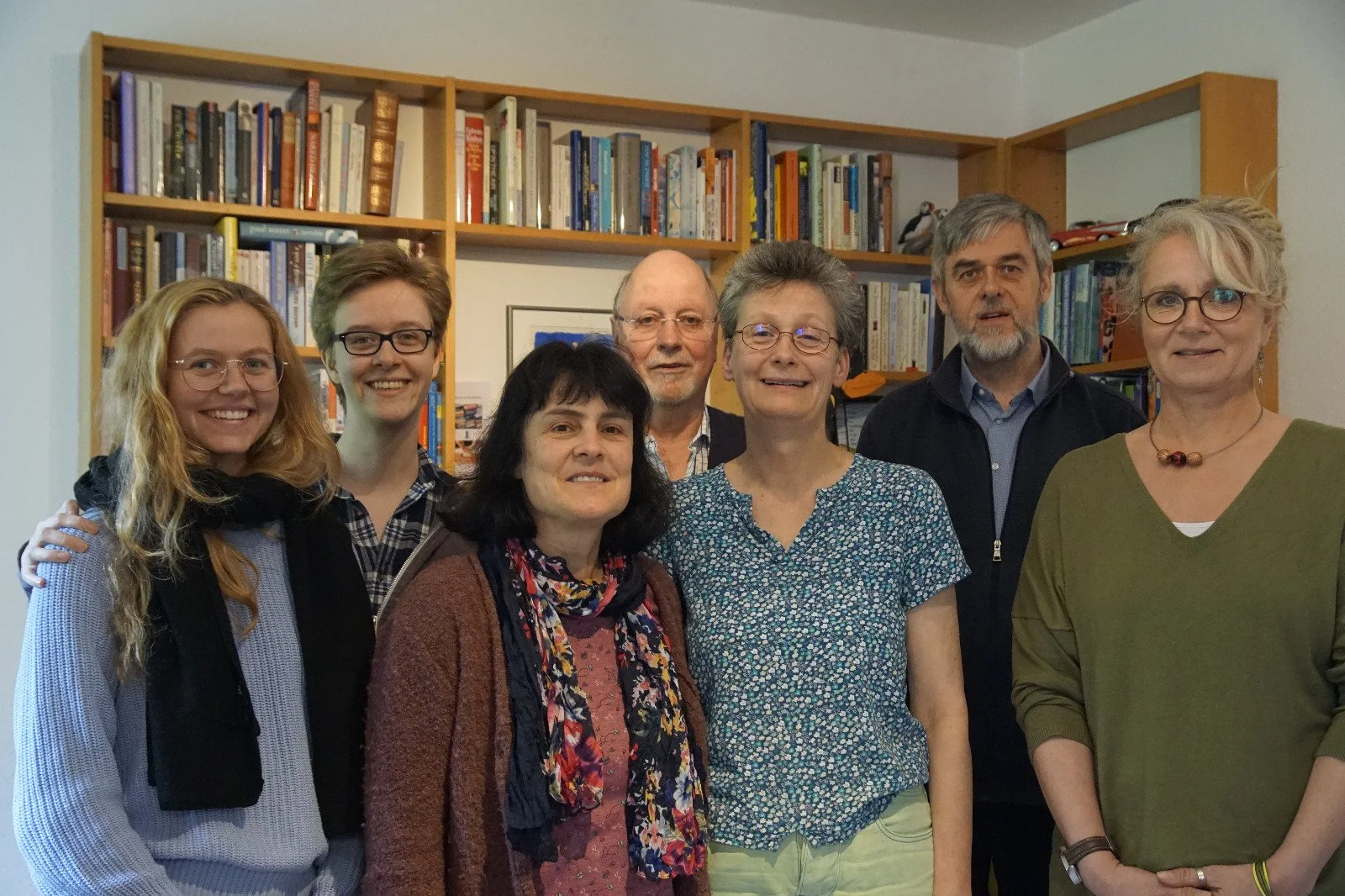 Sieben Personen unterschiedlichen Alters. Vorne fünf Frauen, hinten zwei Männer, die alle in die Kamera lächeln. Hinter ihnen ein großes Bücherregal.