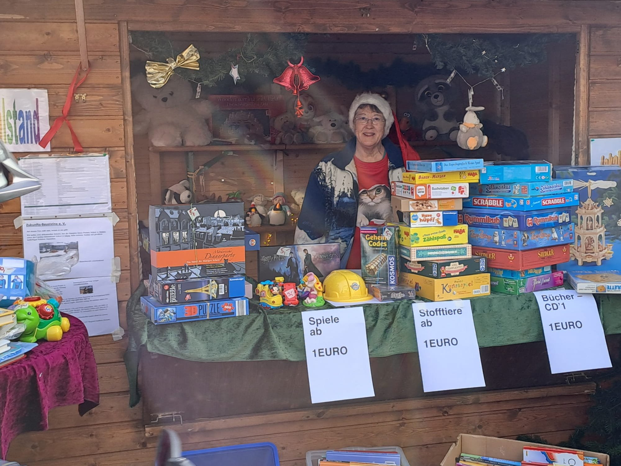 EIn Stand auf einem Weihnachtsmarkt. Eine Verkäuferin, die in die Kamera lächelt. Vor ihr verschiedene Spiele, Plüschtiere etc. mit Zettel mit der Aufschrift "Spiele ab 1 Euro", "Stofftiere ab 1 Euro" etc.