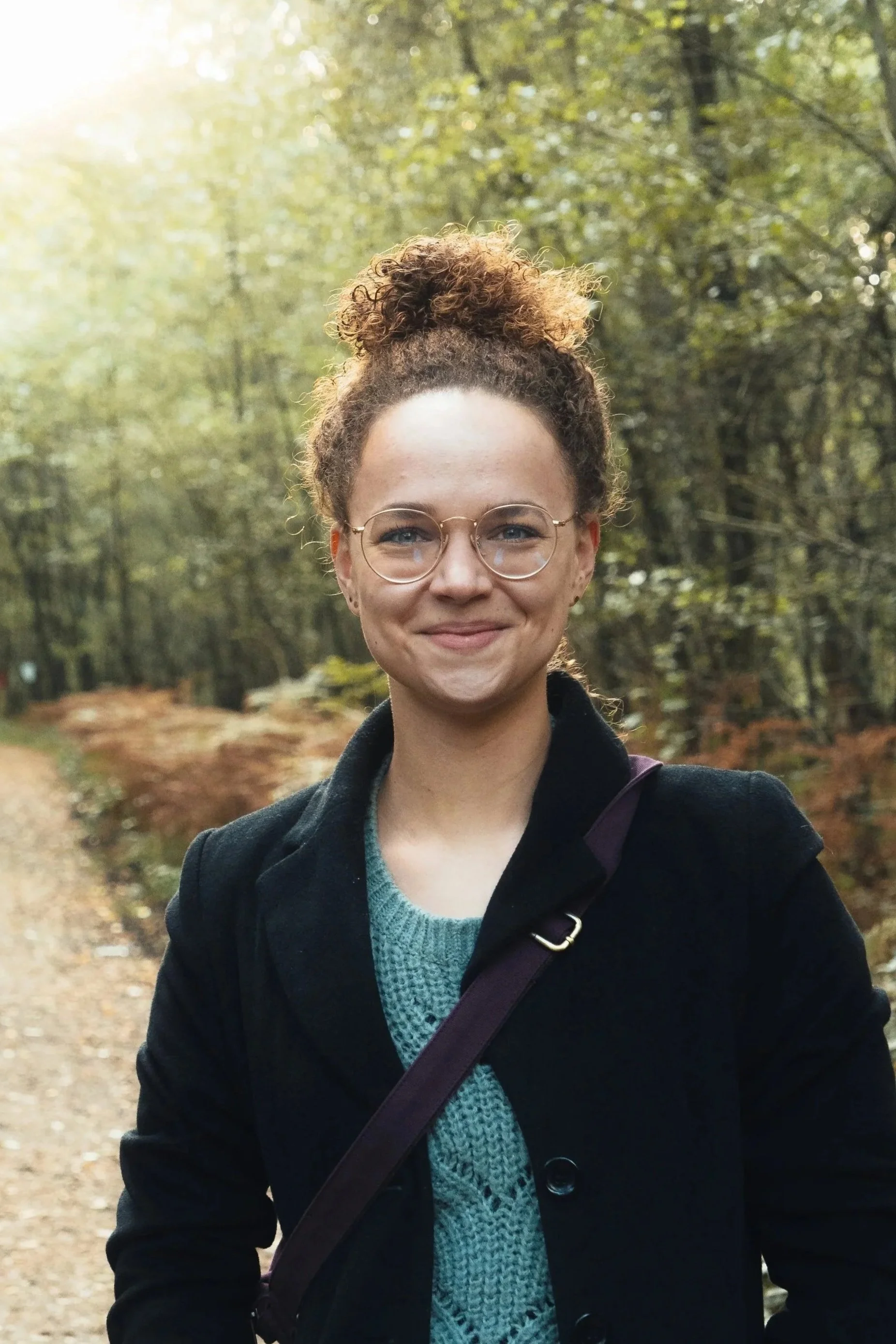 Junge Frau mit Brille und lockigem Haar in einem Wald im Herbst, lächelnd.
