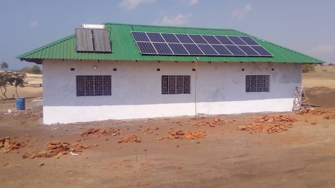 Ein Haus mit grünem Dach und einer Solaranlage darauf. Auf dem Boden liegen Backsteine.