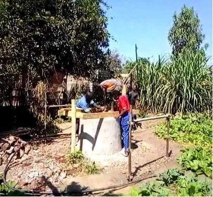 Zwei Personen stehen um einen Trinkwasserbrunnen herum. Um den Brunnen herum befinden sich verschiedene Pflanzen.
