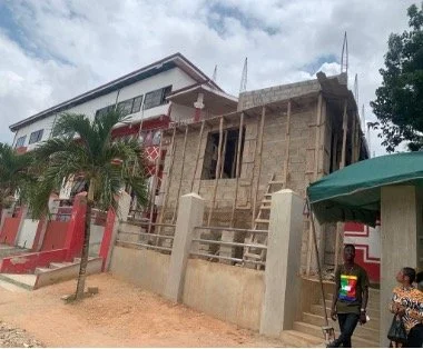 Die Außenmauern eines Gebäudes, im Aufbau, mit Gerüsten. Davor eine Palme. Zwei Personen stehen ebenfalls neben dem Gebäude.