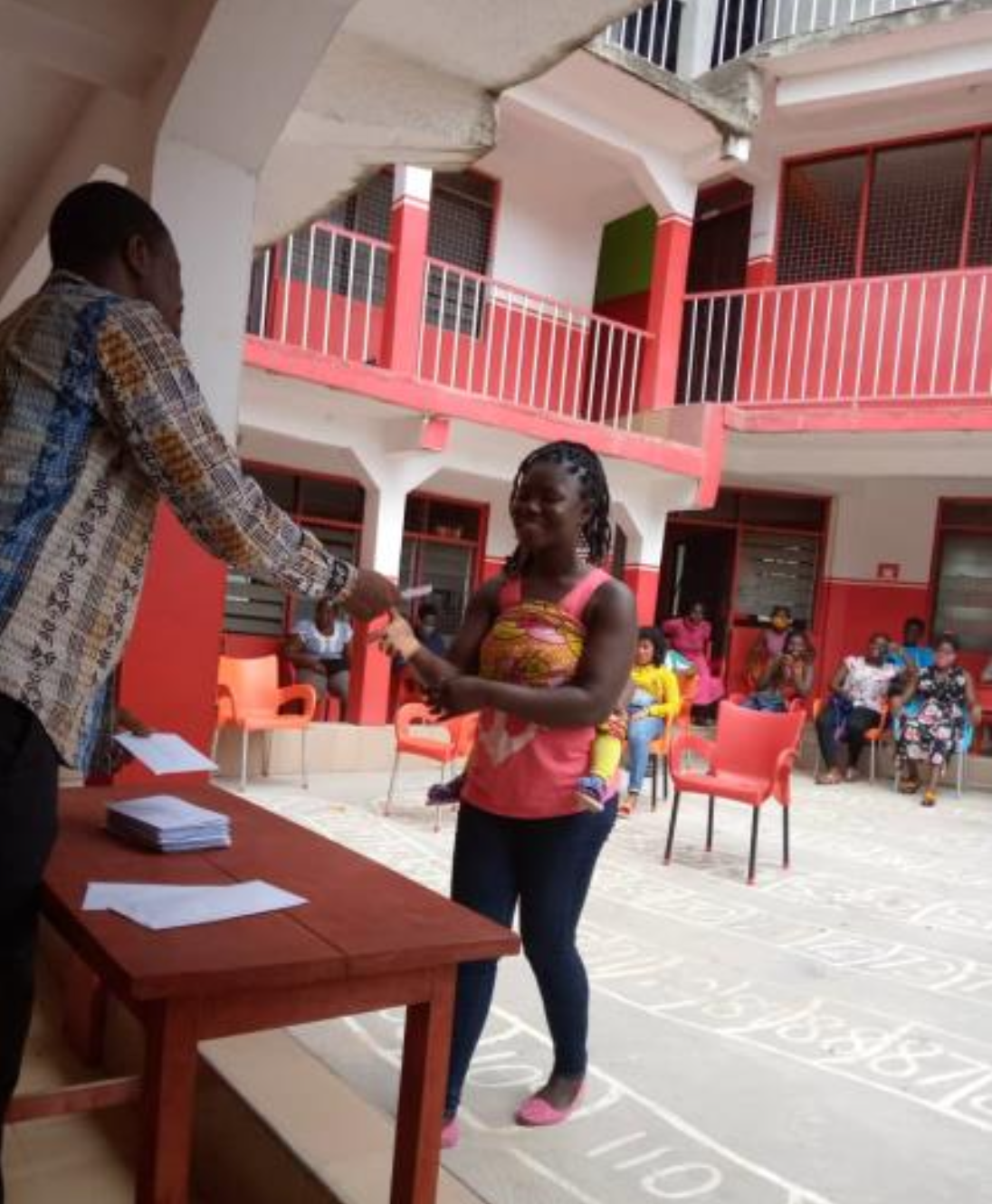 Eine Frau mit Braids nimmt lächelnd etwas entgegen, was ihr ein Mann reicht. Vor dem Mann ein roter Tisch. Beide befinden sie in einem Innenhof. Im Hintergrund sieht man Personen, die auf Stühlen sitzen.
