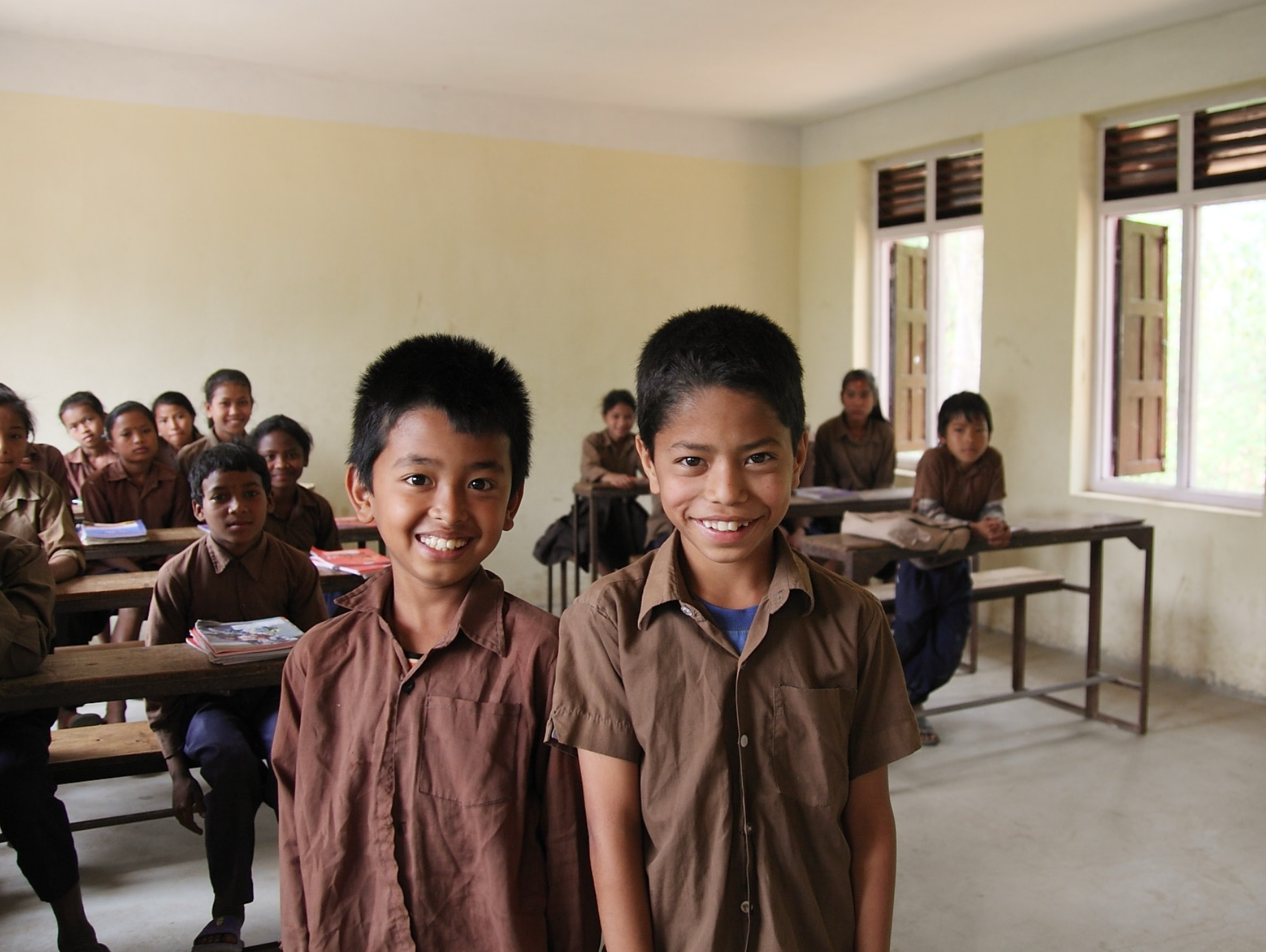Im Vordergrund zwei nepalesische Grundschüler, die in die Kamera lächeln. Hinter ihnen weitere Schüler*innen, die an Tischen in einem Klassenraum sitzen.