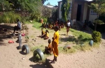 Der Schulhof einer Schule, mit Kindern, die gelbe Gewänder tragen. Aus dem Boden ragen Autoreifen.