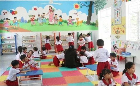 Grundschüler*innen mit rot-weißer Schuluniform in Vietnam in einem Klassenzimmer. Hinten an der Wand ein großes Bild und darunter die Schulbibliothek: mehrere Regale mit zahlreichen Büchern darin.