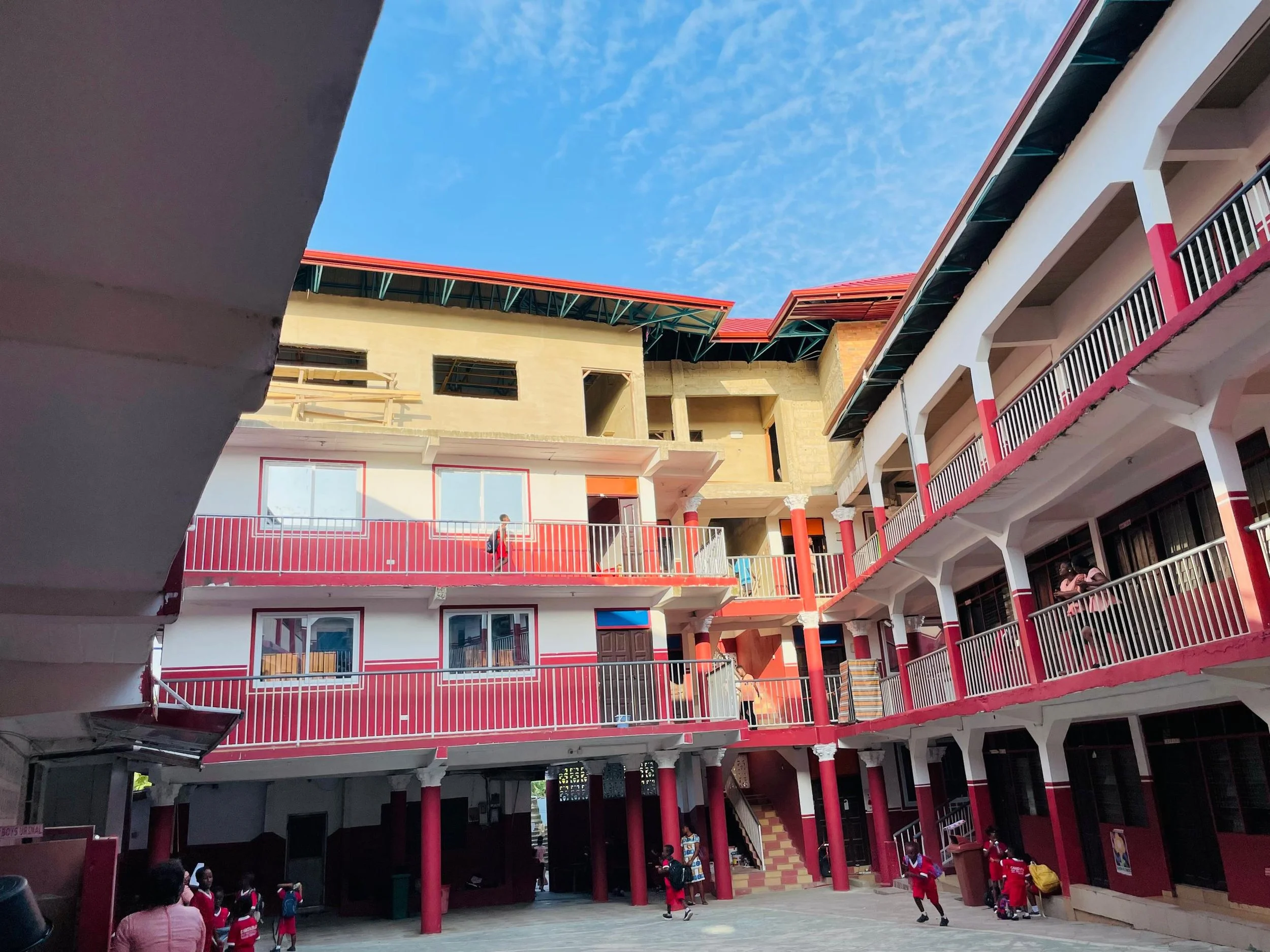 Ein rot-weißes Gebäude mit oberer Etage in gelber Farbe, aus Sicht des Innenhofs. Im Innenhof sind Kinder sichtbar. Am oberen Bildrand der blaue Himmel mit leichten Schleierwolken.