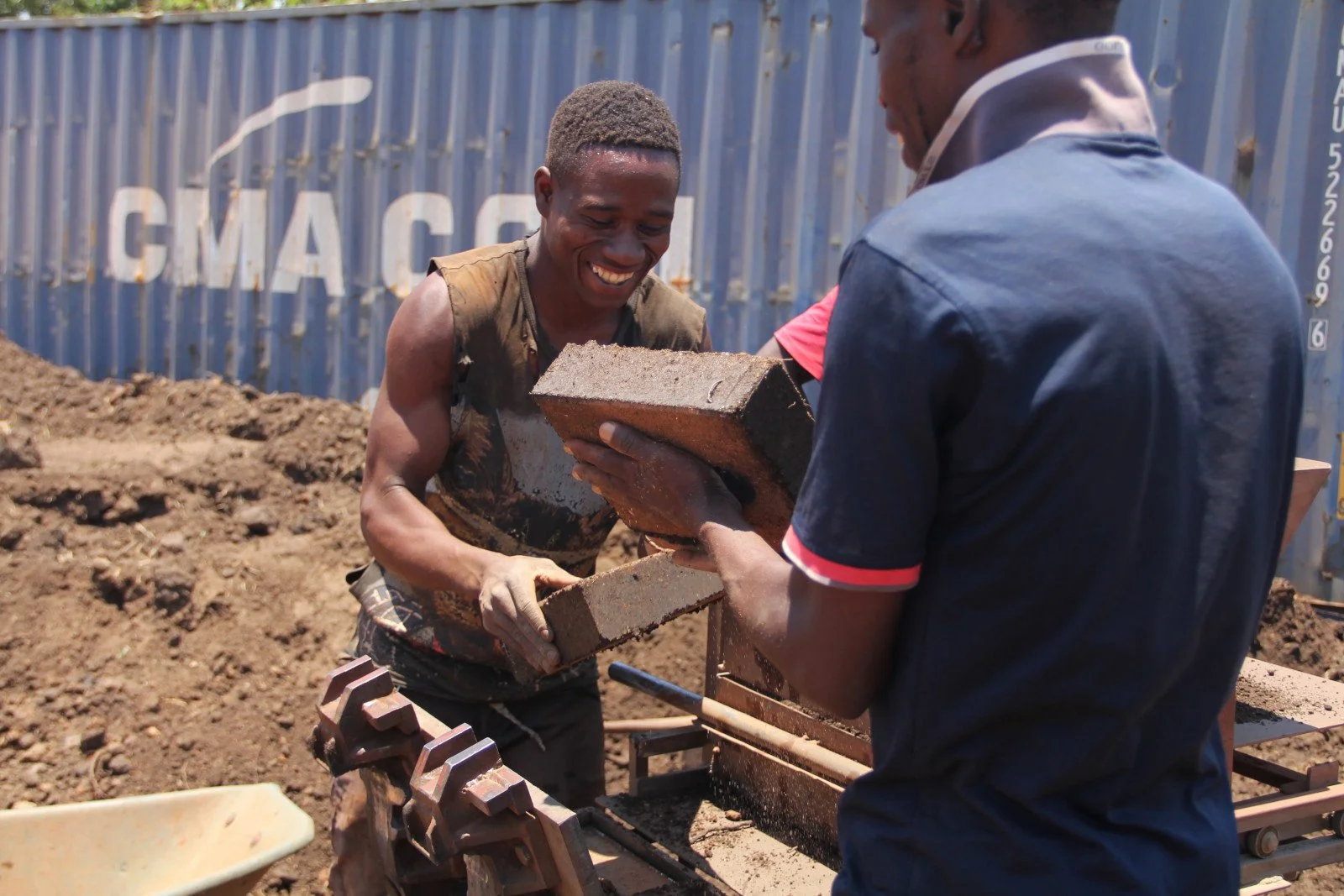Zwei Männer auf einer Baustelle, der eine ist von hinten zu sehen, der andere von vorne lächelnd. Sie heben Mauerziegeln. Copyright: Atmosfair
