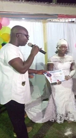 Ein Mann mit Brille spricht in ein Mikrofon. Vor ihm steht eine Frau, mit weißem Puder auf der Haut und einem weißen eleganten Kleid. Der Mann übergibt ihr ein Zertifikat.