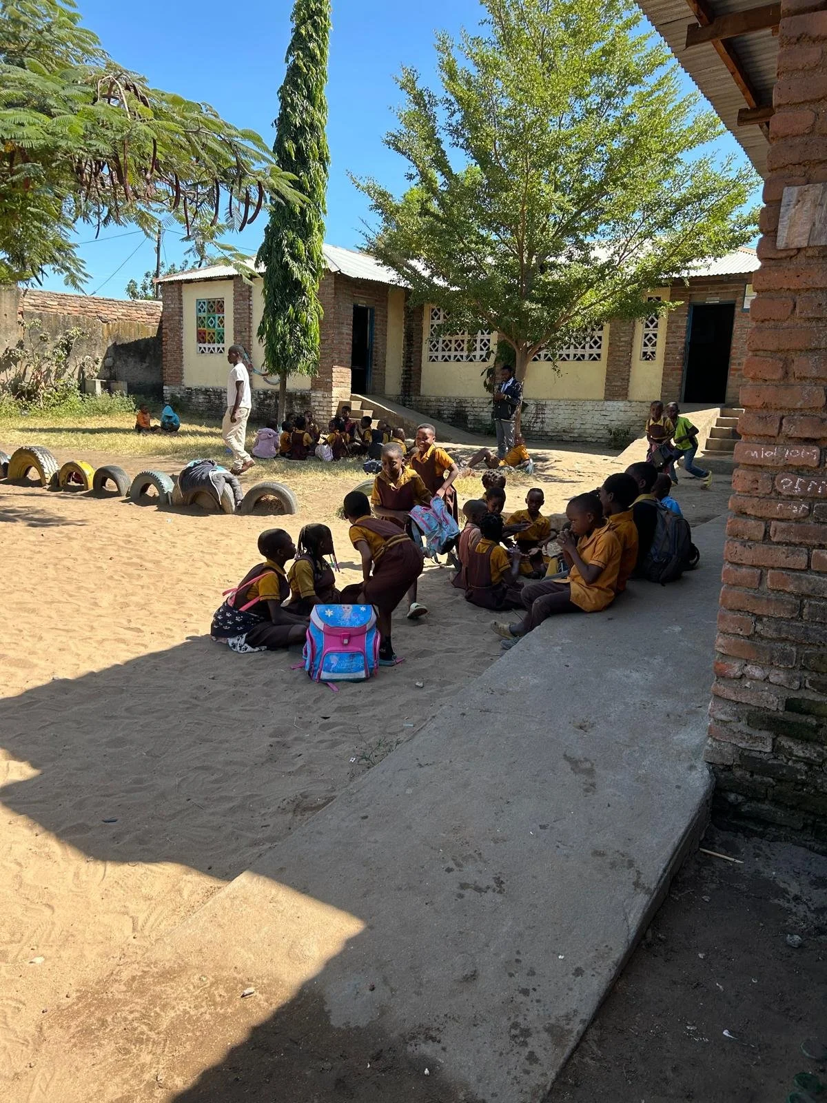 Schulhof in Malawi, Afrika. Im Schatten sitzen Kinder in gelber Schuluniform, neben ihnen ihre Schulranzen. Weiter hinten ein Erwachsener und ein Gebäude. Aus dem sandigen Boden ragen hochkant aufgestellte Autoreifen.