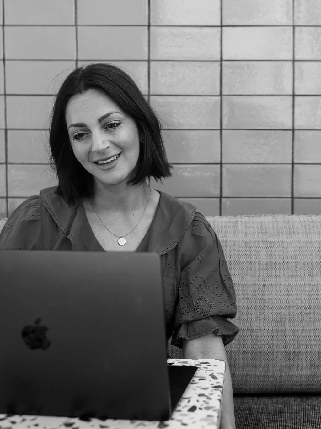 A woman with dark hair sitting at a table with a laptop, smiling, in front of a tiled wall.