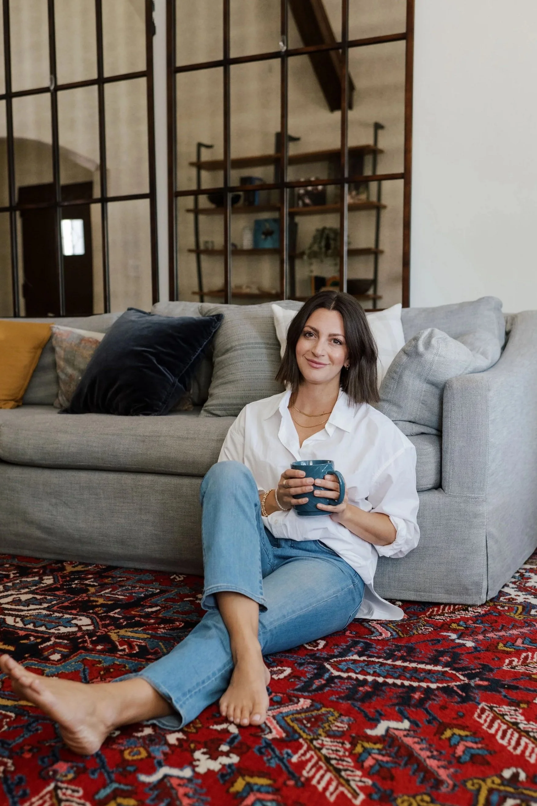 Woman sitting on a colorful patterned rug, leaning against a gray sofa with assorted pillows, holding a blue mug, smiling in a cozy living room.