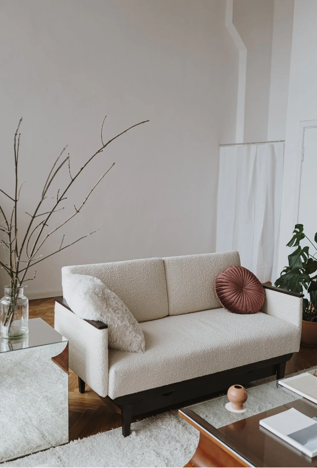 Living room with a white sofa, brown round throw pillow, white fluffy cushion, glass-topped side table with a vase of bare branches, and potted plant in the background.