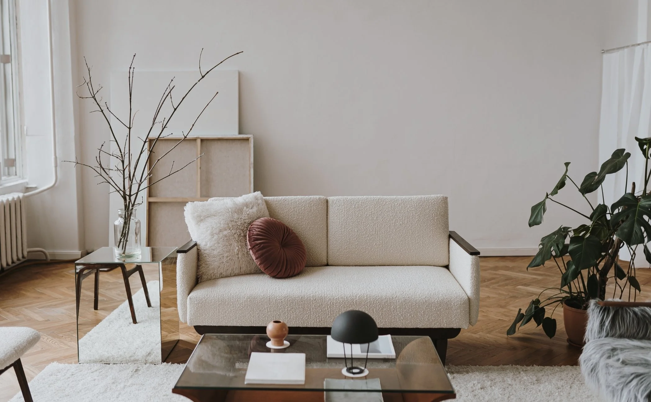 Living room with a white sofa, round cushion, and fluffy pillow, a glass coffee table holding decorative objects, a side table with a vase and branches, large window, and potted plants.