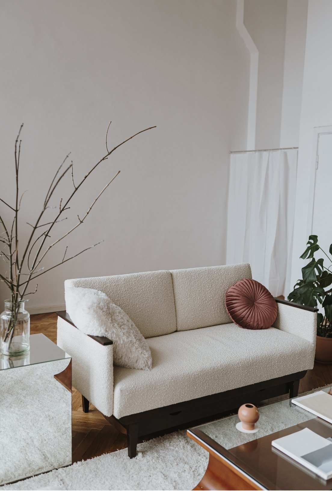 A beige sofa with a fluffy white pillow and round brown pillow inside a living room, next to a glass coffee table and a side table with a glass vase and dried branches, and a potted plant in the corner.