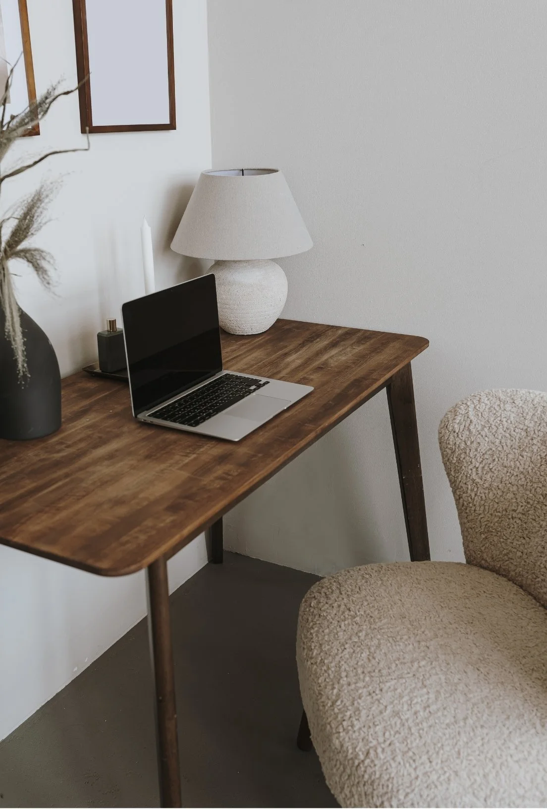 A wooden desk with a laptop, a white lamp, a black vase with dried flowers, and a candle in a holder. Part of a beige, textured chair is visible.