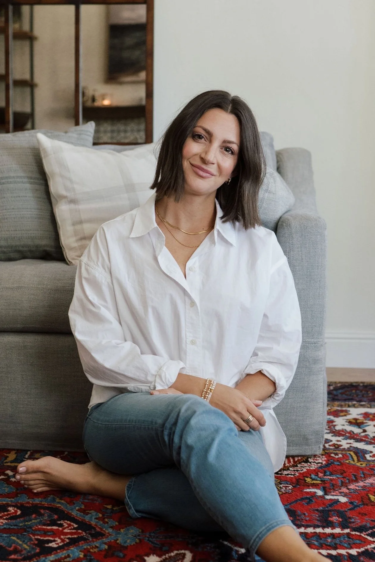 A woman with shoulder-length brown hair, wearing a white button-up shirt and jeans, sitting cross-legged on a red patterned rug in a cozy living room. She is smiling at the camera, with pillows and a sofa behind her.