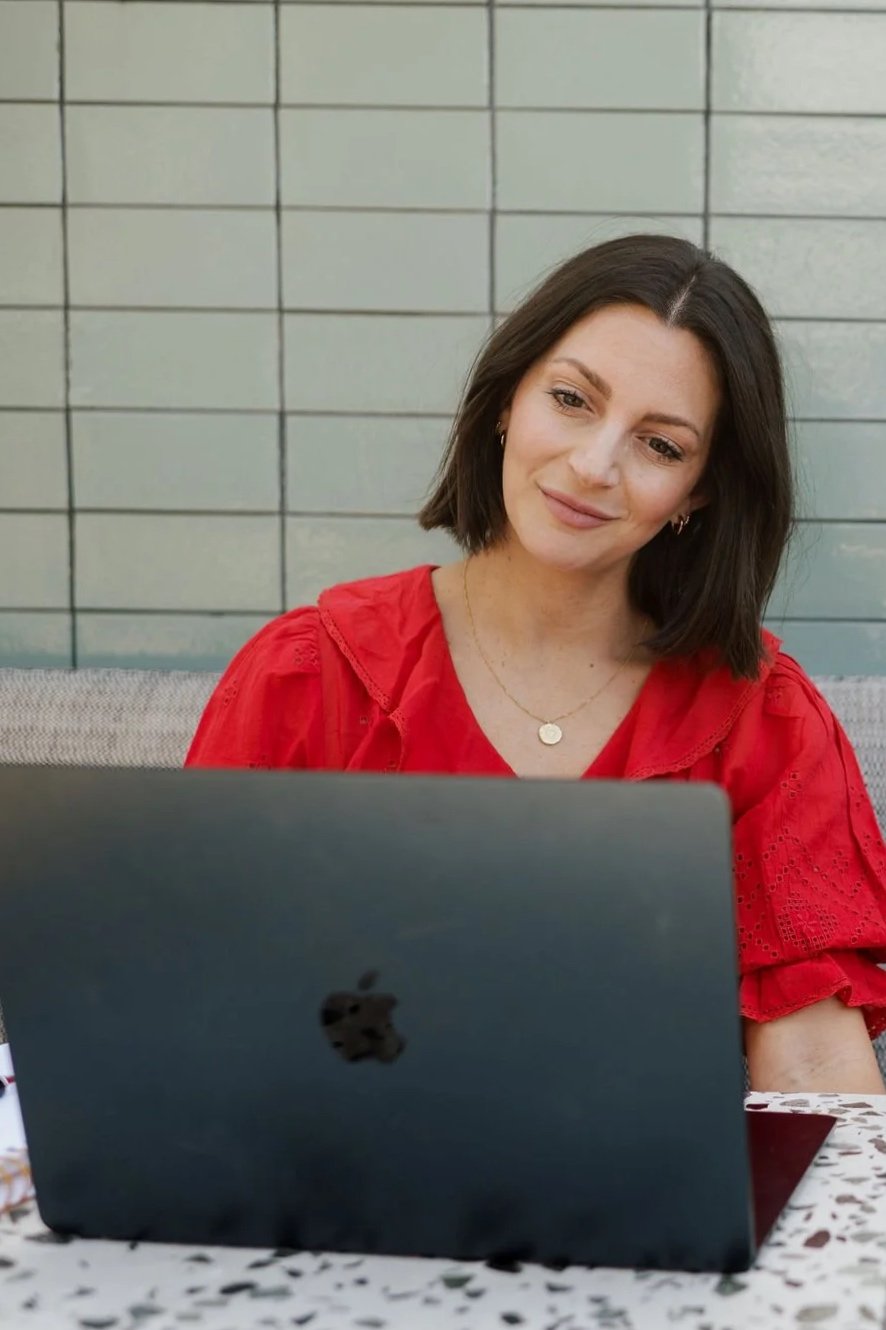 A woman with shoulder-length dark hair sitting outdoors in front of a laptop, wearing a red blouse and a gold necklace.