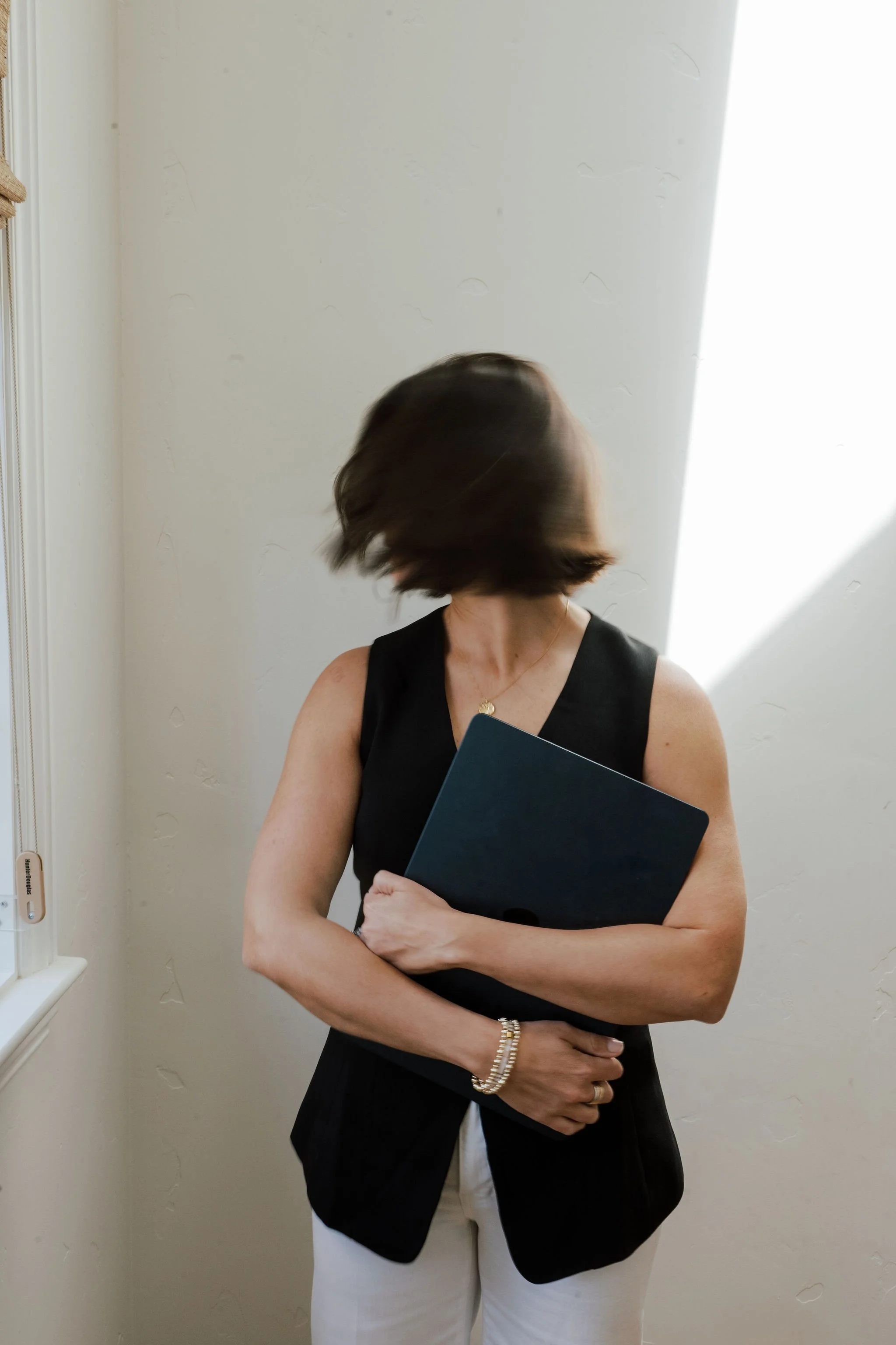 Woman in black sleeveless top holding a dark blue folder with hair covering face, standing in front of a beige wall.