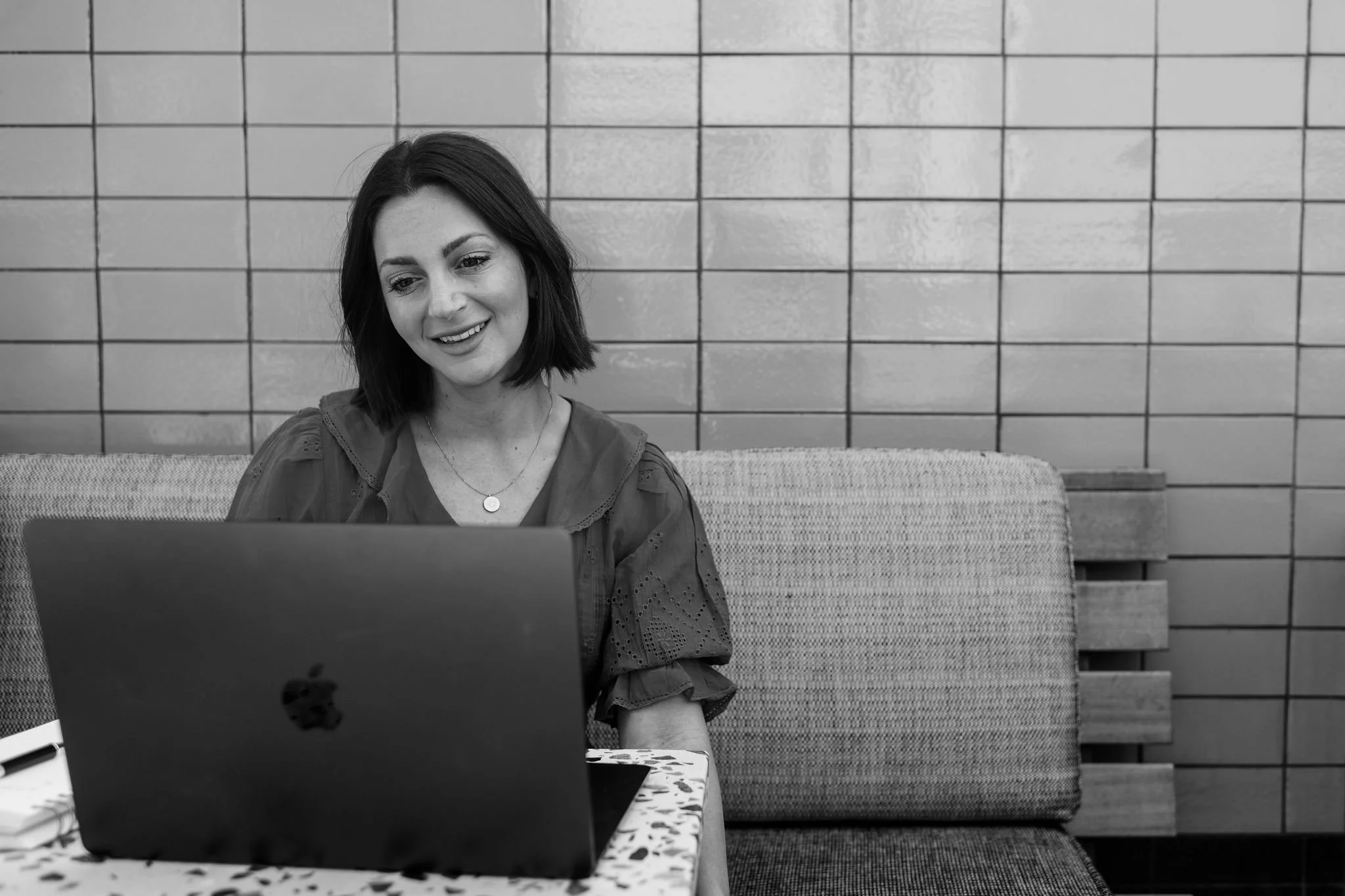 A woman with shoulder-length dark hair sitting at a table, smiling and looking at a laptop in front of her, in a room with tiled walls.