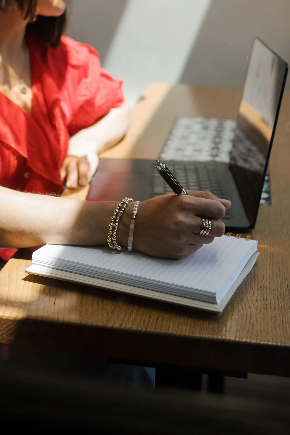 A person's hand is writing in a notebook at a wooden desk, in front of a laptop. The desk has sunlight and shadow patterns. The person is wearing bracelets and rings.