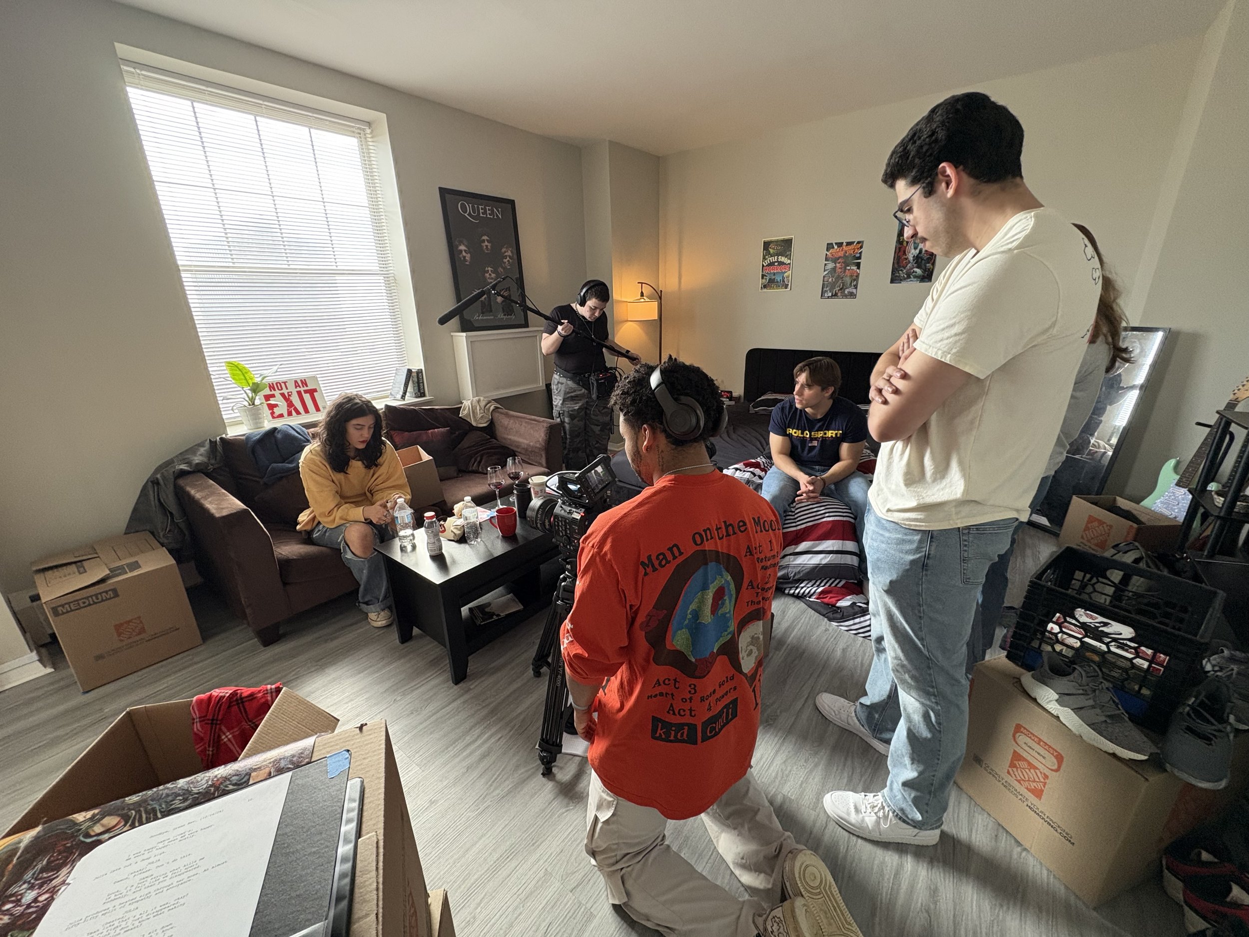 Filming crew setting up for a scene in a living room with several people, including a woman sitting on a sofa, a man in a white shirt and glasses, and a person with headphones operating a camera, with a person standing by a window and a woman holding