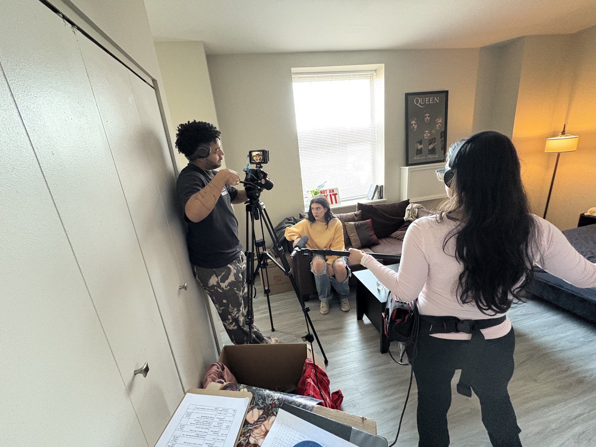 A woman being interviewed while sitting on a couch in a living room, with a man filming her and a woman holding a microphone.