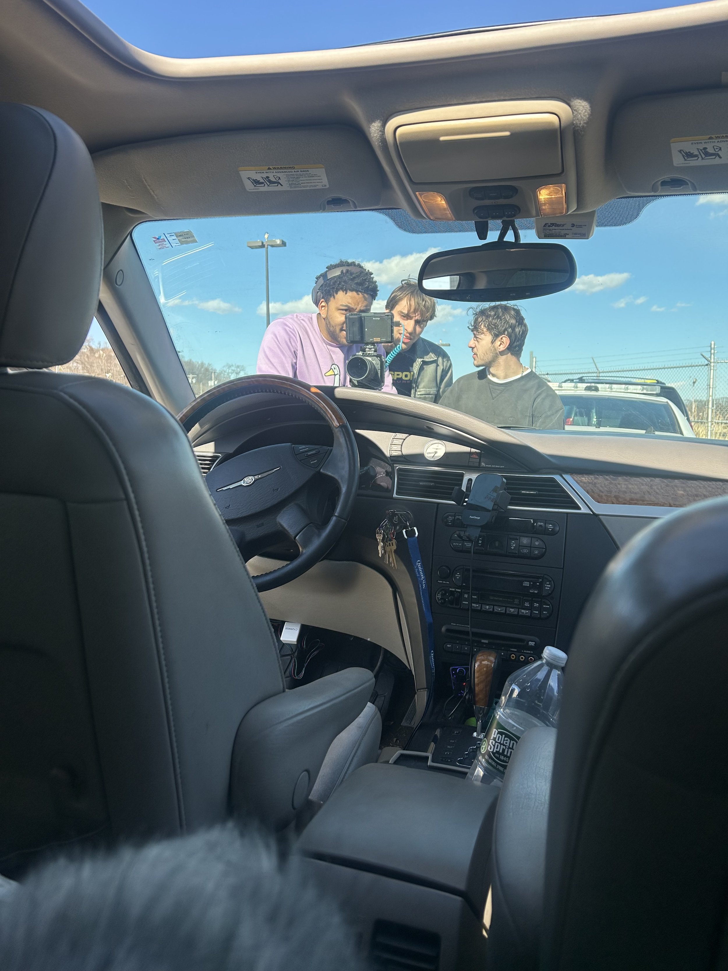 View from inside a car shows three people outside, looking in through the windshield, with a clear blue sky and parking lot in the background.