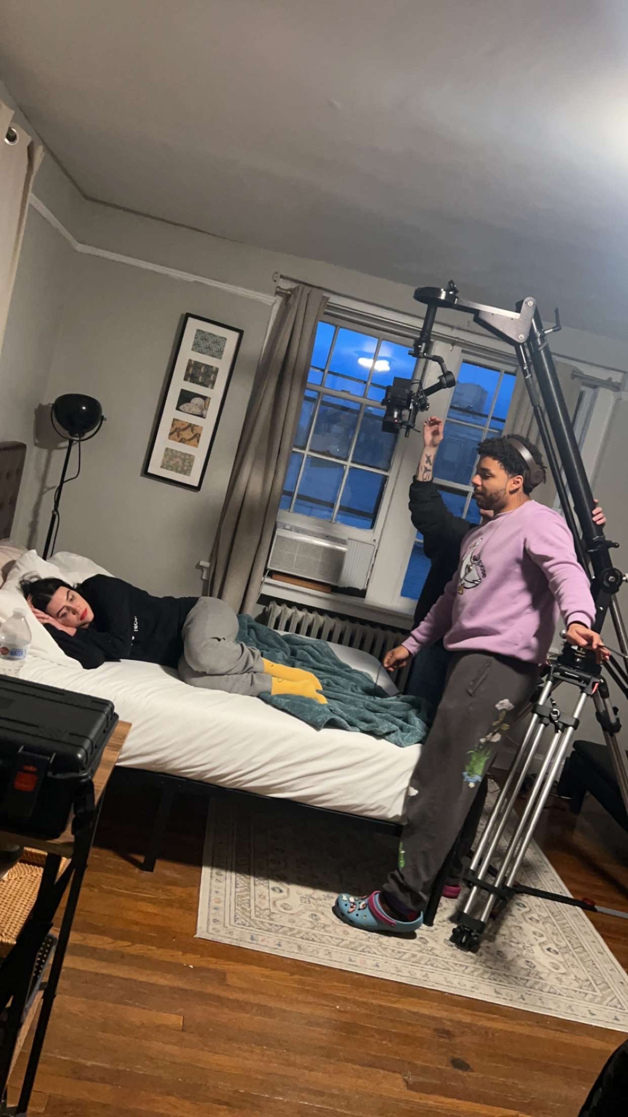 A woman lying on a bed with her head resting on her hand, wearing a black shirt and grey sweatpants, looking at a man operating camera equipment in a bedroom with a window, curtains, framed artwork, and wooden floors.