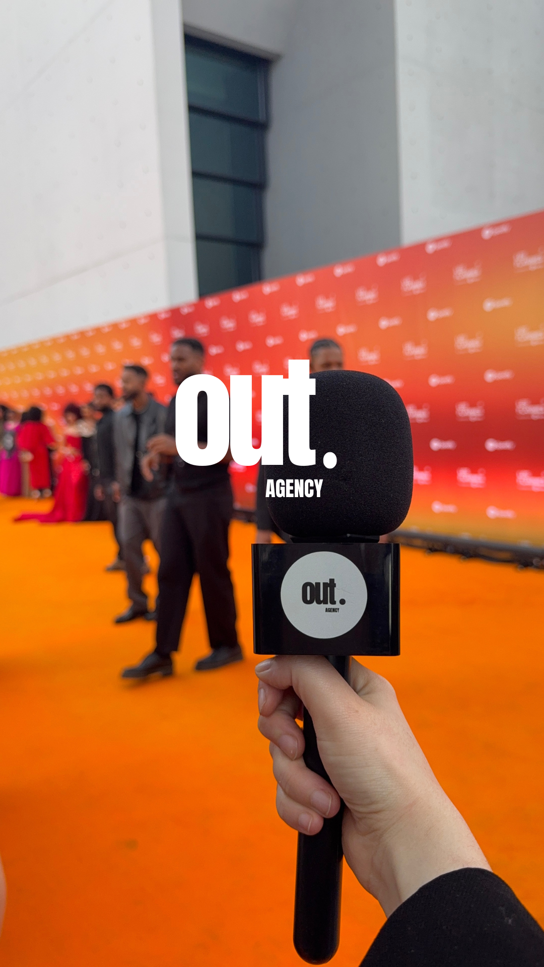 Une main tient un microphone noir devant un groupe de personnes en ligne sur un fond orange avec le texte 'out. AGENCY'.