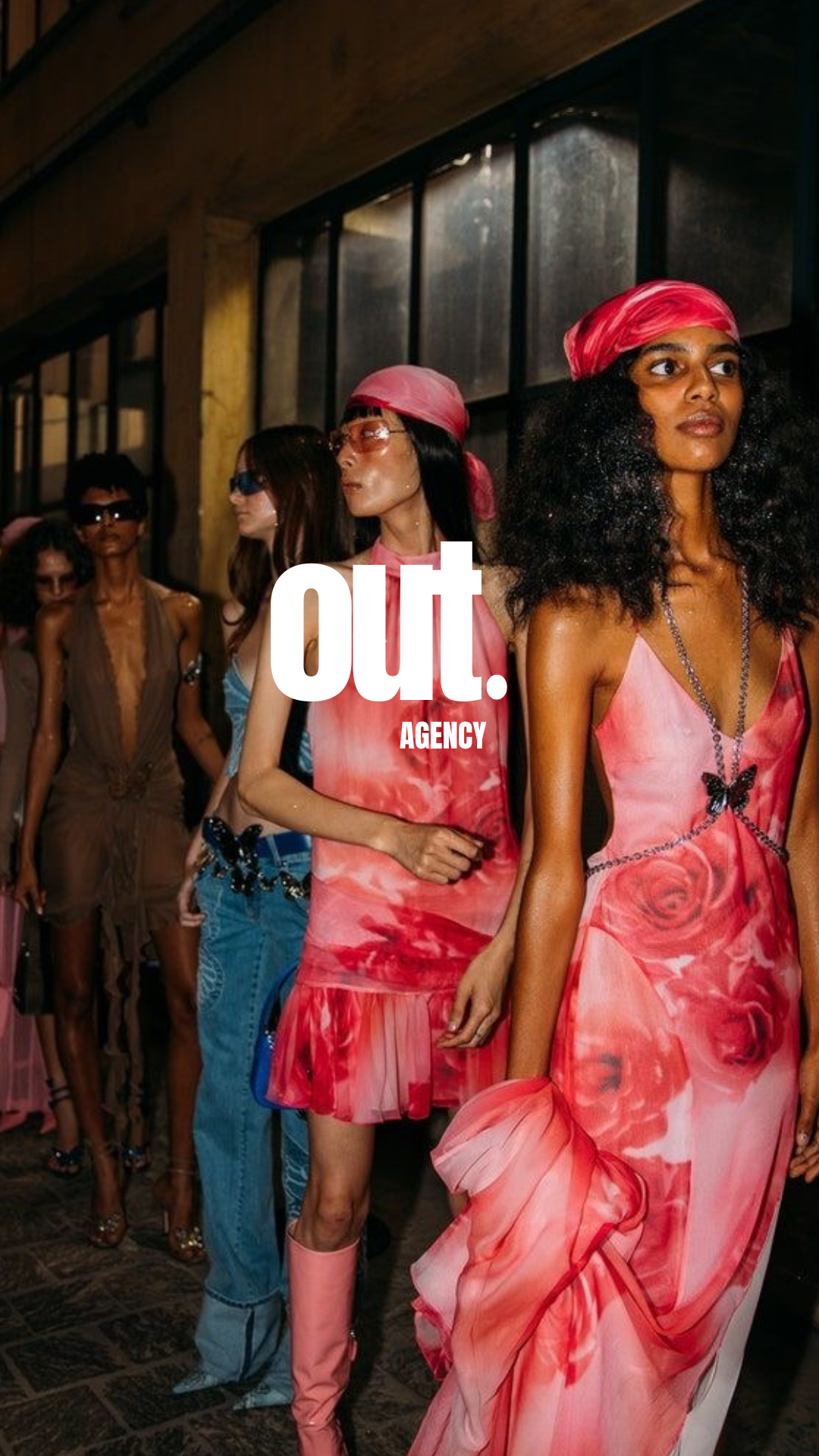 Modèles féminins marchant dans un défilé de mode avec vêtements colorés, notamment des robes imprimées roses et rouges, portant des accessoires comme des chapeaux, des lunettes de soleil, et des bijoux, dans un espace industriel ou artistique.