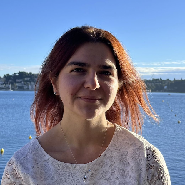 A young woman with long brown hair standing outdoors in a mountainous area with green vegetation and clear skies.