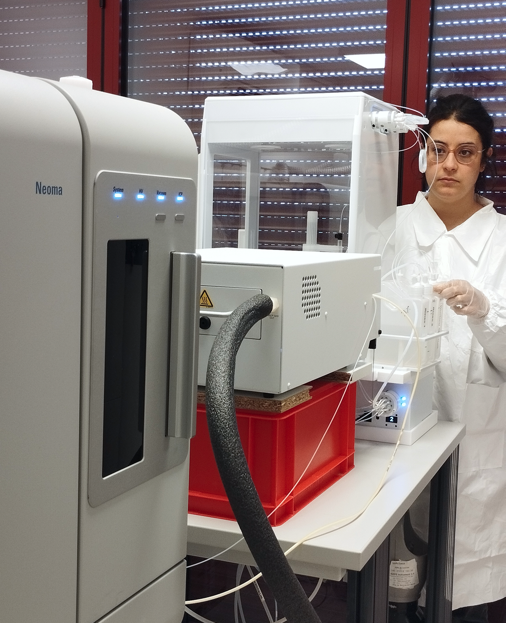 A scientist in a white lab coat and gloves working with laboratory equipment, including a Neoma sterile environment system and a controlled chamber, in a laboratory with blinds covering the windows.