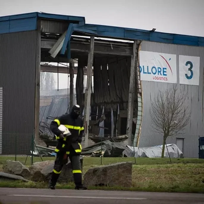 Damaged industrial building with a large hole in the side, a firefighter walking in the foreground, and a small tree on the right.