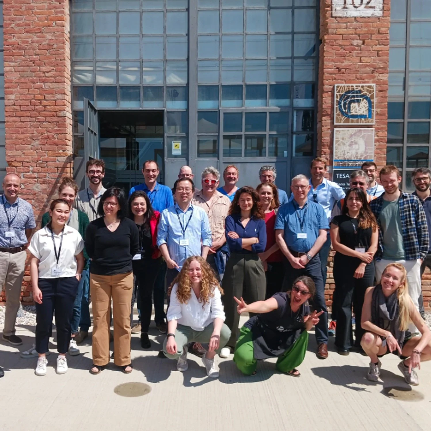 A large group of people posing together outside a building with large glass windows and brick walls. They are smiling and appear to be attending a professional or academic event, with some wearing name tags.