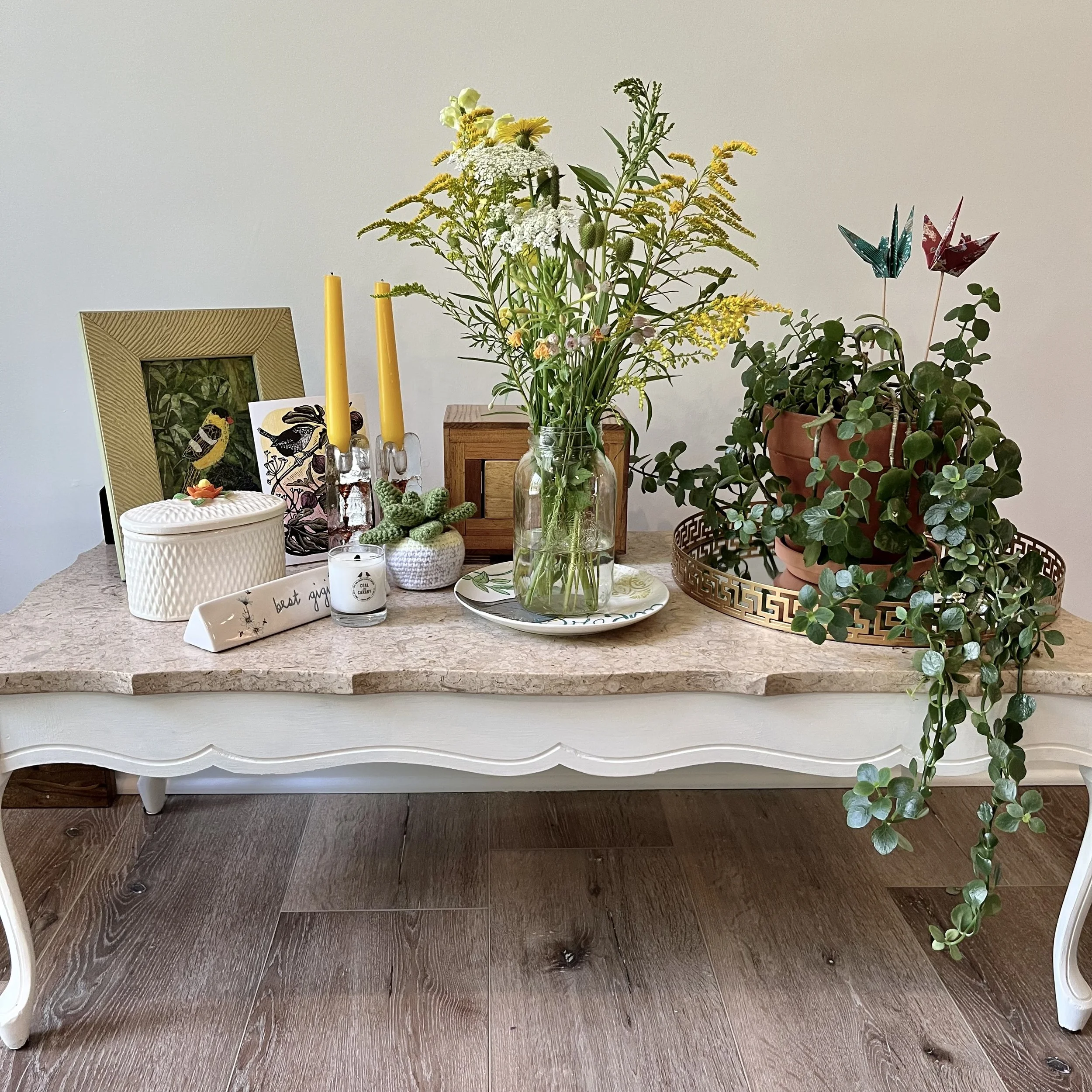 Table with marble top, collection of plants, small wooden box, green bird picture.