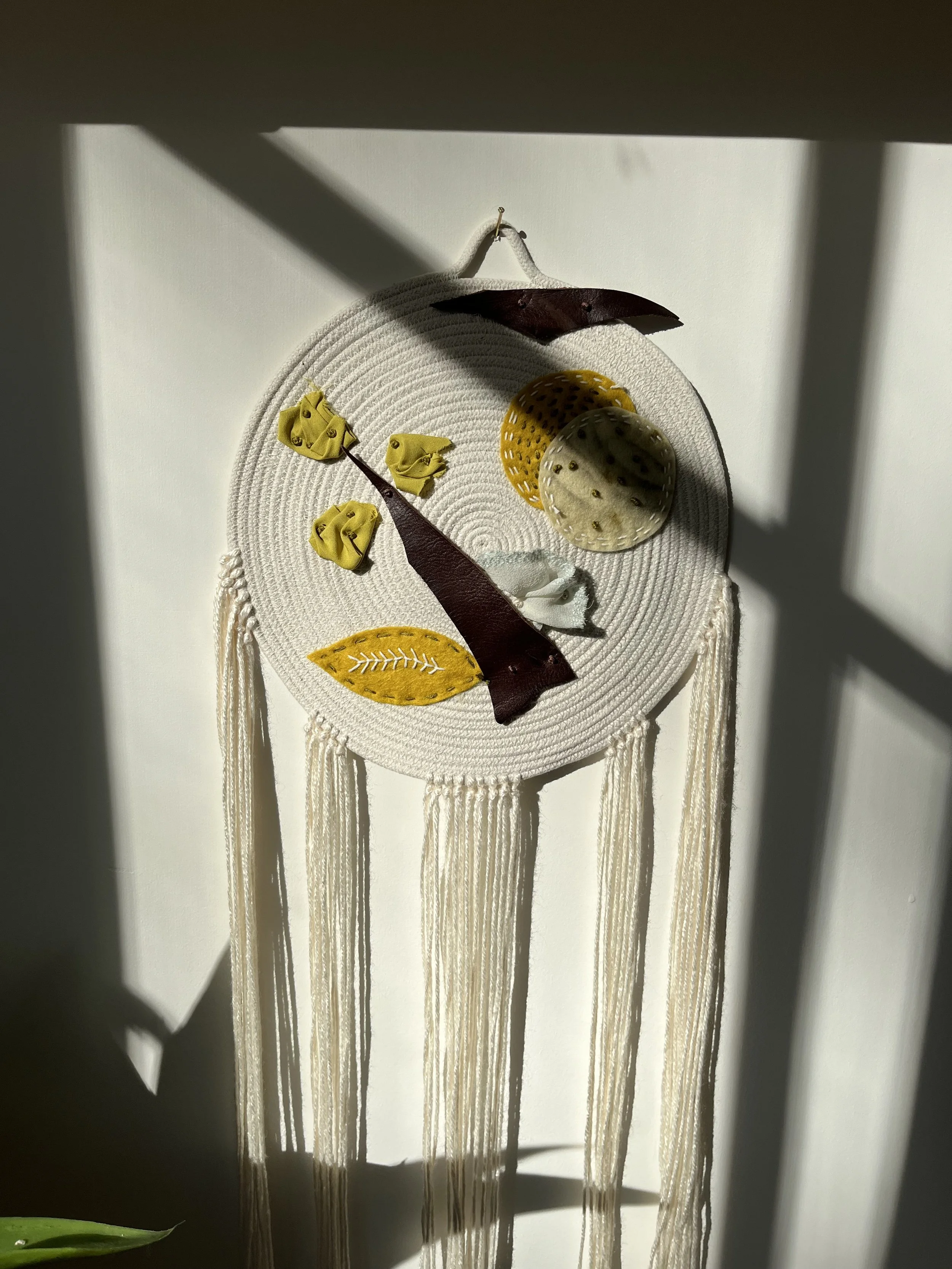 Decorative rope wall hanging featuring  hanging tassels, adorned with felt leaves and geometric shapes in yellow, brown, and white. 