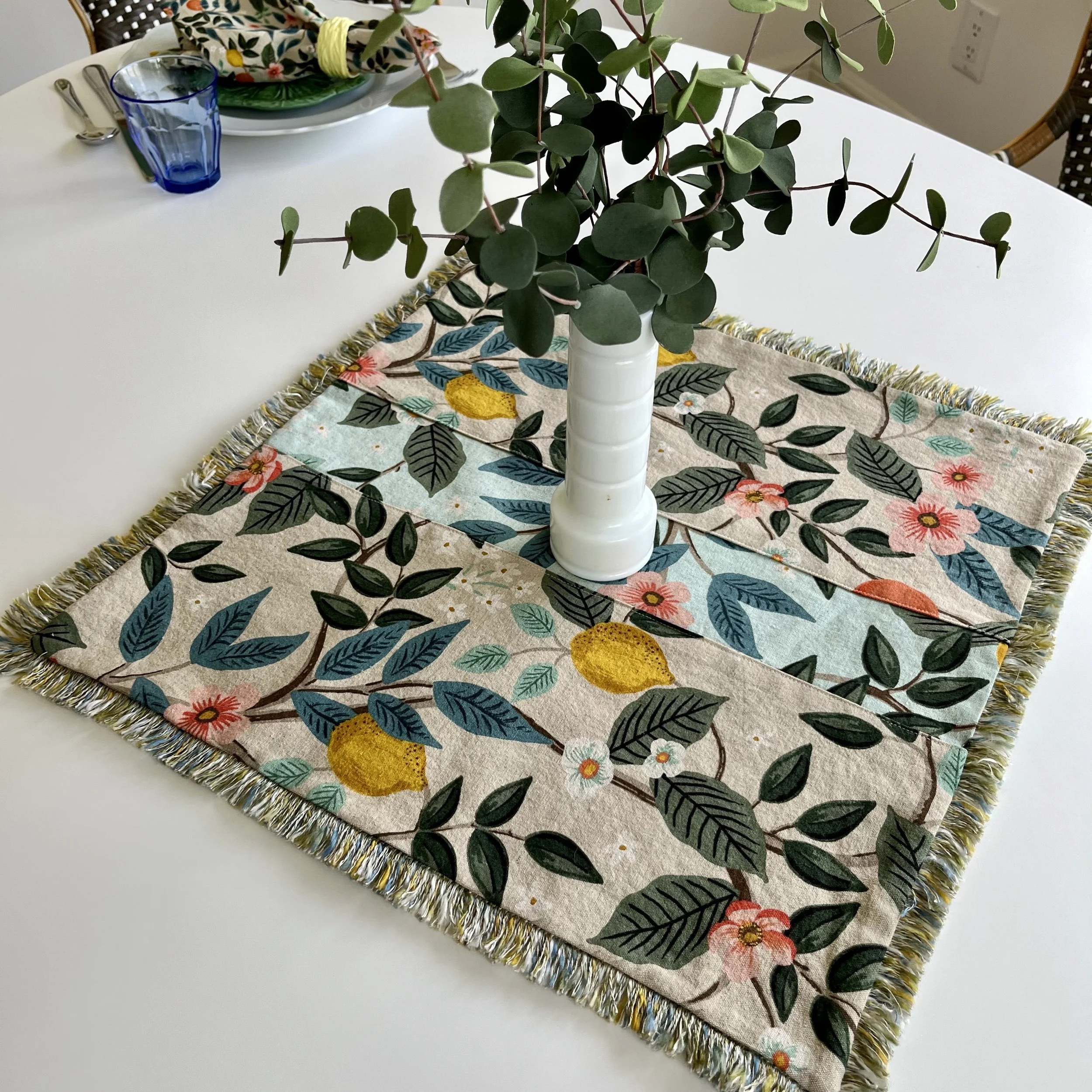 A white table with a colorful small square floral and citurs table runner, a white vase with green foliage, and a place setting with a blue glass, plates, and napkins.