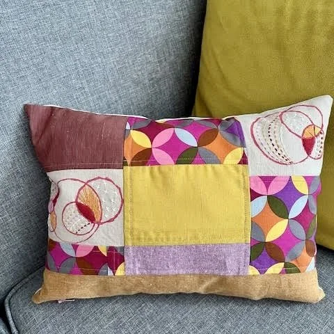 A pieced cotton linen decorative toss pillow with hand printed circular designs and hand stitching. In shades of purple and golds.