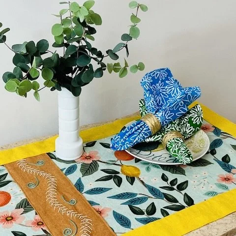 A table with a colorful floral / citrus pattern table runner, with a yellow linen border.