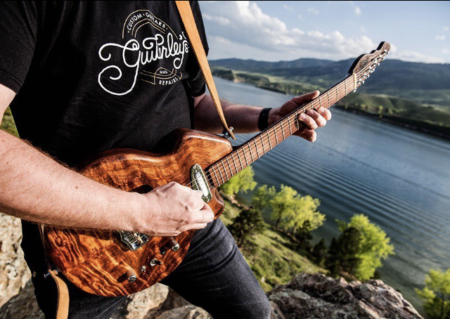 Person playing an electric guitar outdoors near a lake with trees and hills in the background.
