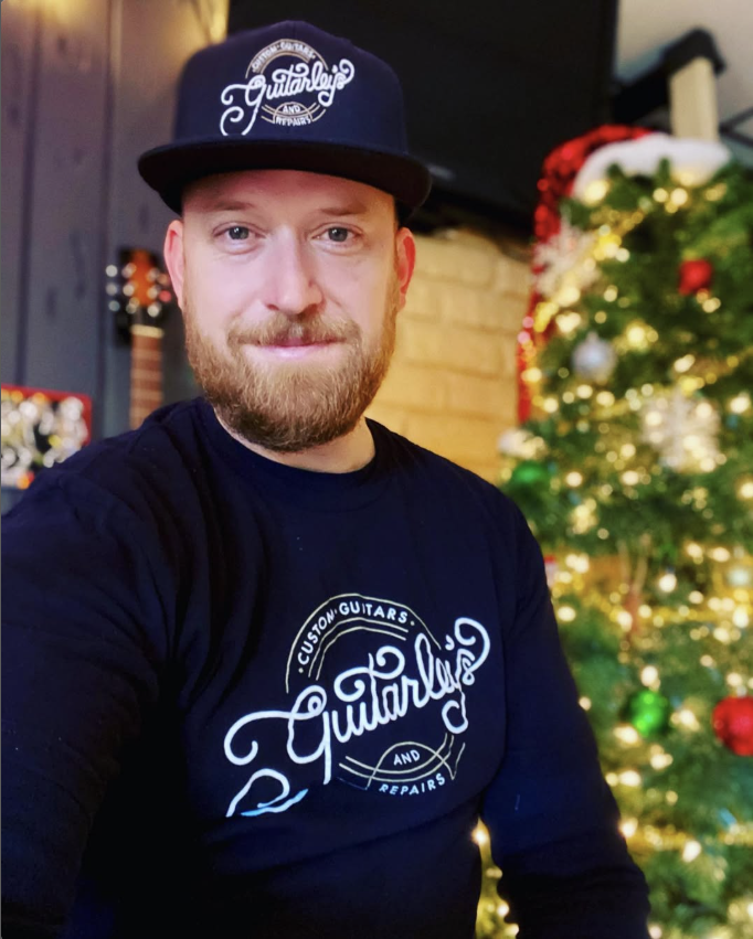 A man with a beard wearing a black baseball cap and a black shirt with a guitar-related logo, sitting in front of a decorated Christmas tree with ornaments and lights.