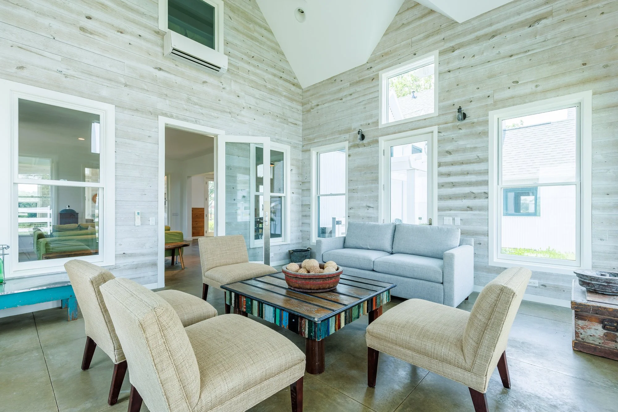 Interior real estate photography of a living room featuring tons of windows, polished concrete floors, cathedral ceilings, and sitting area. Walls are made of shiplapped white oak stained real wood, and feature stunning angular design elements