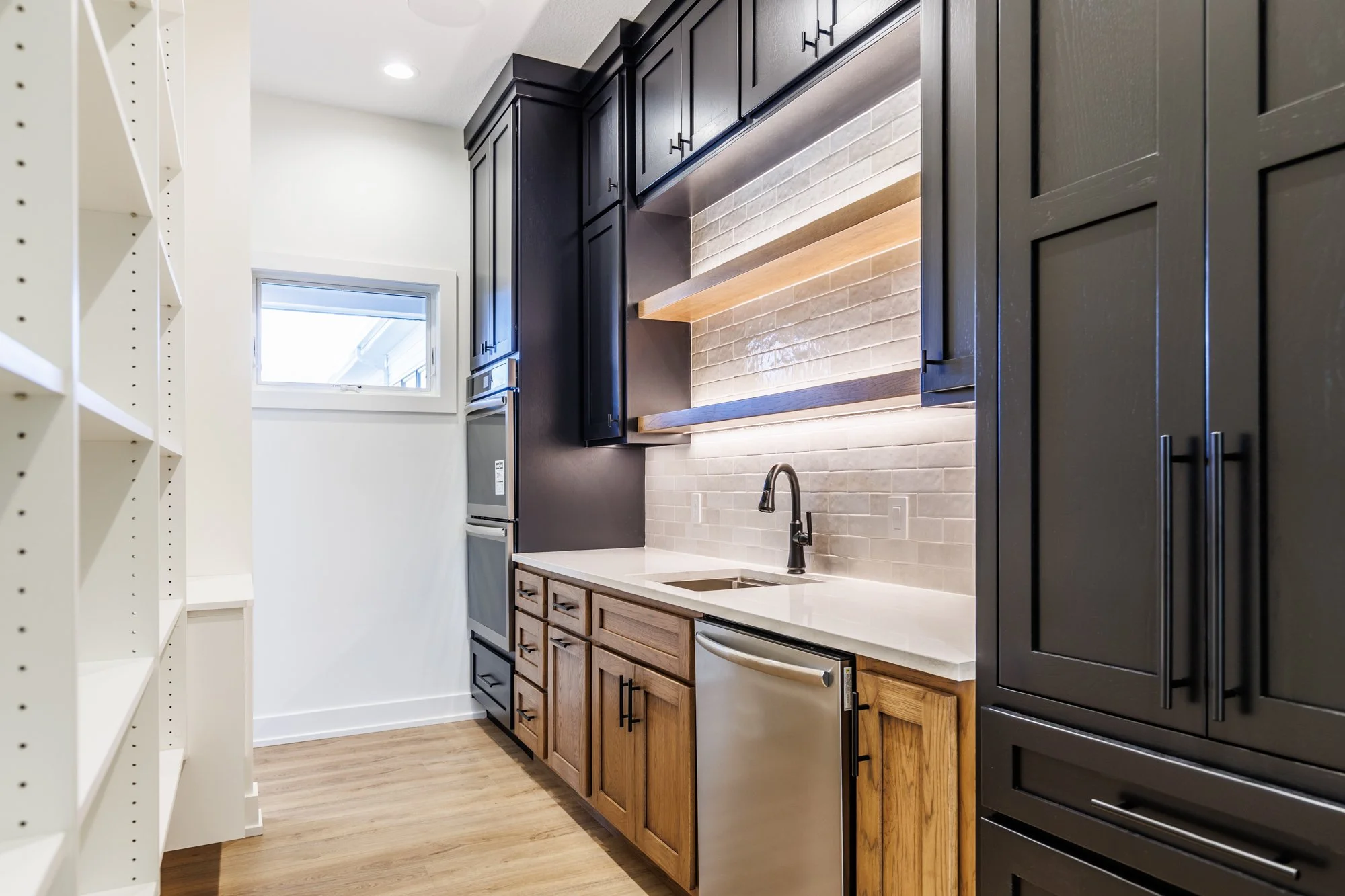 Interior real estate photograph of a butlers pantry. This pantry features plenty of storage, both open and cabinetry, as well as under lit open shelving above the gorgeous sink. Also features additional dishwasher, beautiful countertops, stacked ston