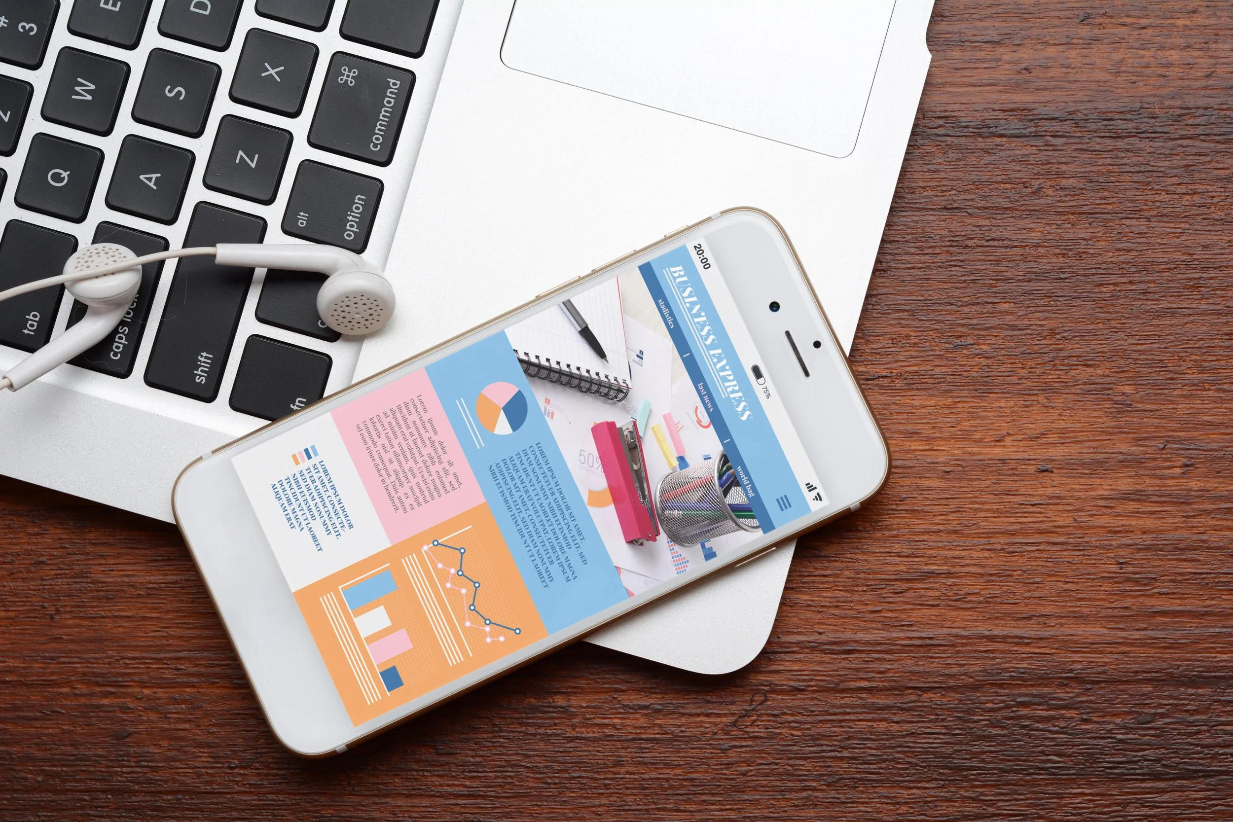 A desk scene with a silver laptop, white earphones resting on the keyboard, a smartphone displaying a business infographic, placed on a wooden surface.