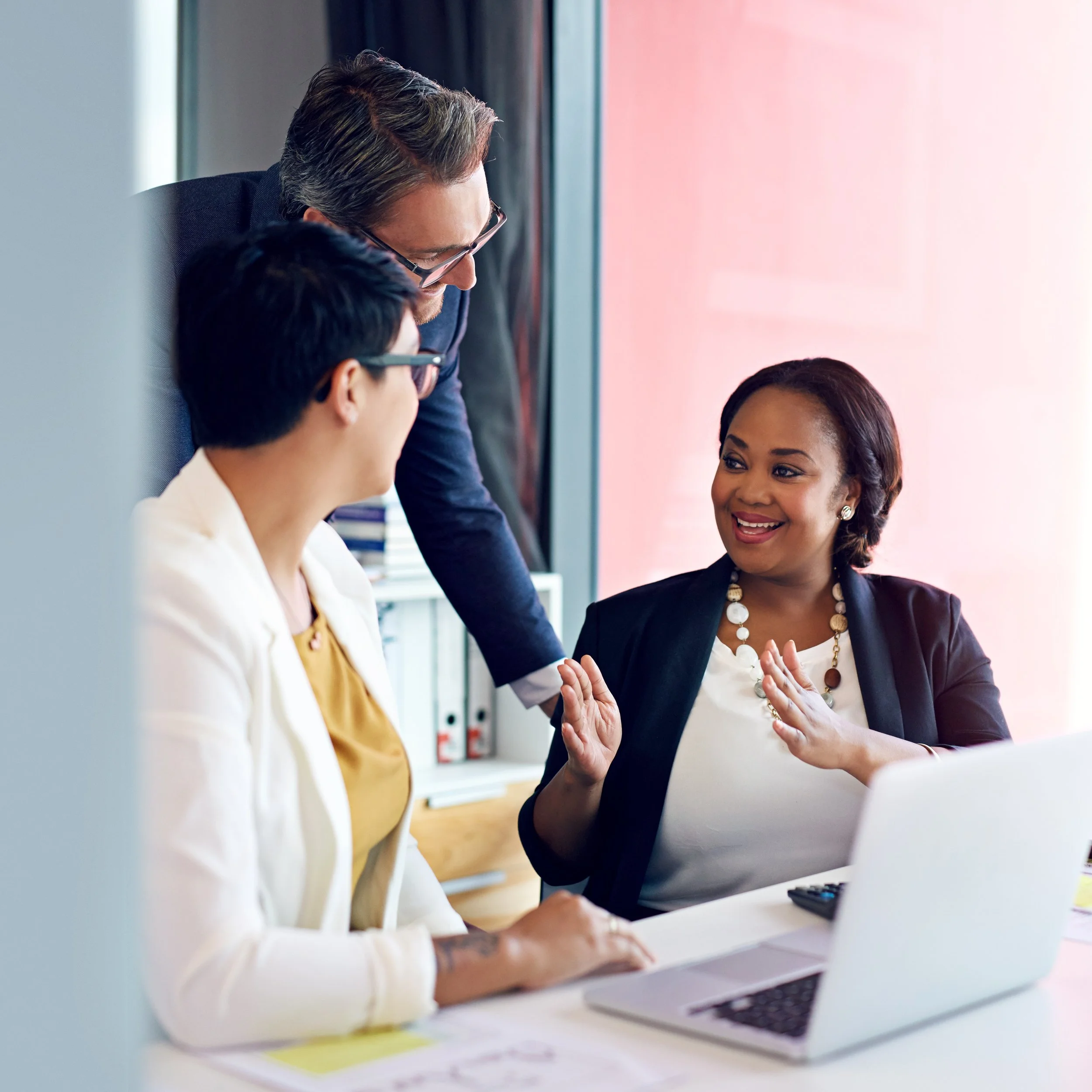 Three business professionals engaged in a discussion in a modern office, with one woman smiling and raising her hand.